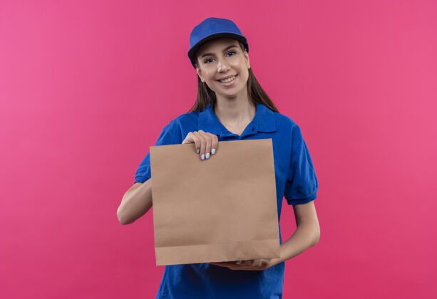 站着身穿蓝色制服 头戴鸭舌帽的年轻送货女孩拿着纸包看着相机友好地微笑着看着女孩包装