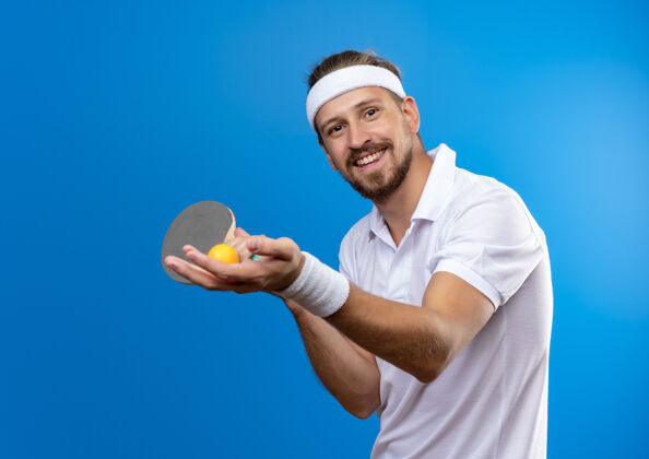 年轻微笑的年轻帅气的运动型男子戴着头带和腕带拿着乒乓球拍 球被隔离在蓝色的空间里腕带抱着头带