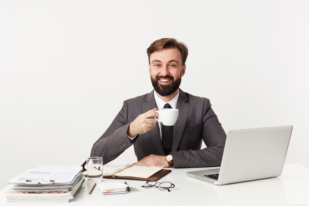 发型一张室内照片 一个留着黑胡子的积极的年轻人 在工作日过得很愉快 他坐在白色墙壁上的桌子旁 在休息时间端着一杯咖啡 面带微笑深色咖啡男性