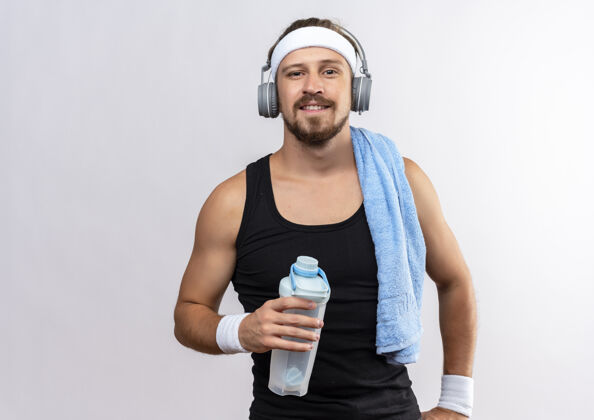 瓶子面带微笑的年轻帅气的运动型男子戴着头带和腕带 头戴耳机 肩上拿着毛巾的水瓶 隔离在白色的空间里水运动帅气
