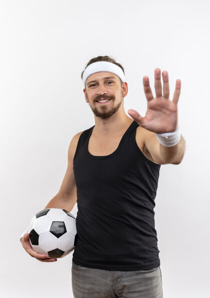 年轻面带微笑的年轻英俊的运动型男子戴着头带和腕带拿着足球 伸出手孤立在白色的空间里足球头带微笑