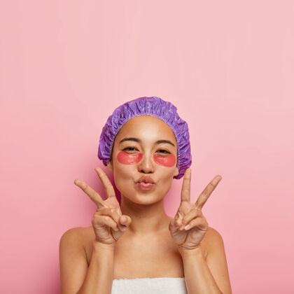 折叠美丽和年轻化的概念美丽的韩国女人做和平的手势 保持嘴唇折叠 脸上有眼罩 戴紫色浴帽 洗澡后享受水疗程序护理情感眉毛