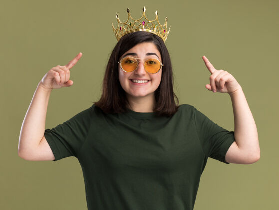 皇冠微笑着的年轻漂亮的高加索女孩戴着太阳眼镜 头顶朝上 两只手孤立在橄榄绿的墙上 留着复印空间橄榄眼镜太阳