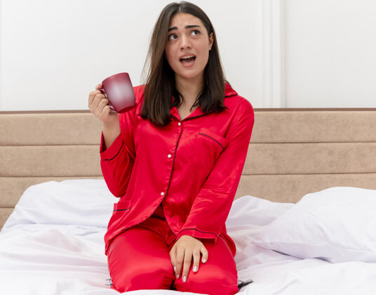 床穿着红色睡衣的年轻美女坐在床上 端着一杯咖啡 抬头望去 在卧室的室内灯光背景下迷茫漂亮看咖啡