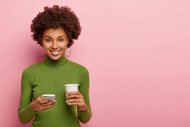 模特表情满意的美女 拿着手机和咖啡走 穿着绿衣服 发短信 在网上聊天交流 隔着粉色的墙孤立卷发美式套头衫