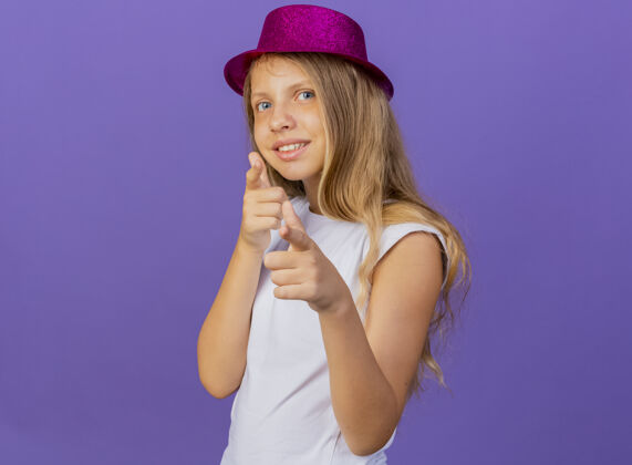 微笑戴着节日礼帽的漂亮小女孩用食指指着镜头开心而积极地笑着 生日派对的概念站在紫色的背景上漂亮正面相机