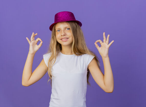 漂亮戴着节日礼帽的漂亮小女孩看着镜头笑眯眯的脸上露出oksing 生日派对概念站在紫色背景上快乐好生日