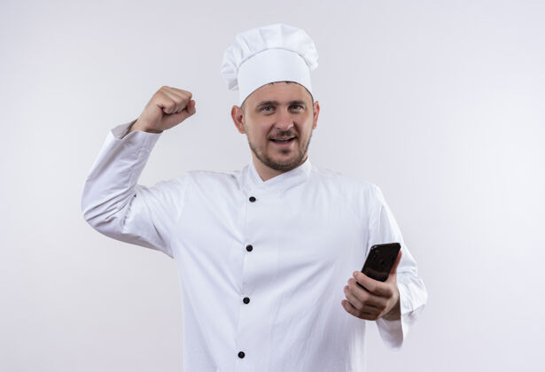 帅气面带微笑的年轻帅哥厨师身着厨师制服手持手机做着坚强的手势隔离在空白处强壮握着厨师
