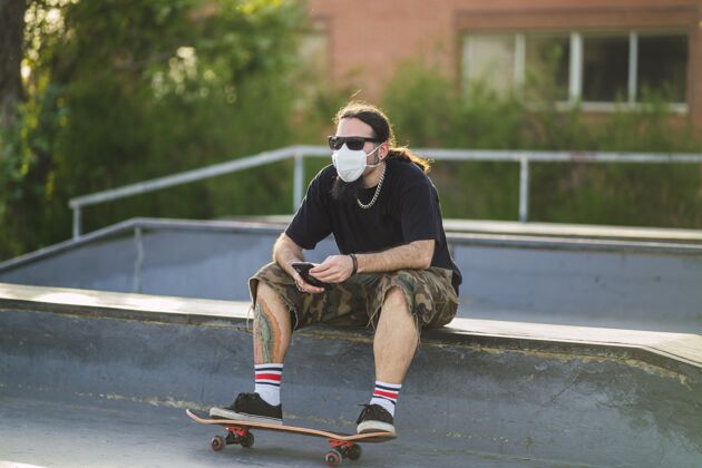 城市戴着医用面罩坐在公园里玩滑板的年轻男性-covid-19概念休闲行动滑板