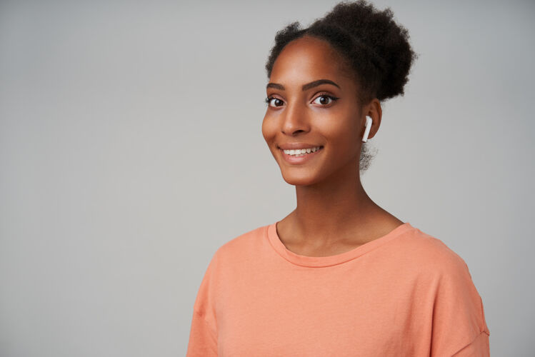 欢快年轻的棕色眼睛 深色皮肤 浅棕色头发的女人的肖像 带着灿烂的微笑 快乐地看着 孤立的灰色衬衫穿着发髻