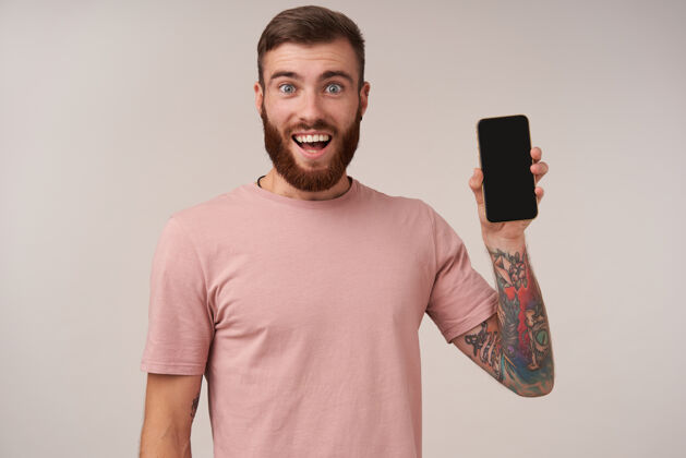 表情快乐的蓝眼睛美丽的胡须 纹身的男性穿着米色t恤 在白色的脸上摆姿势 开心地微笑着 举起手 里面有智能手机无胡子衣服男士