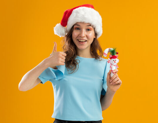 顶身穿蓝色上衣 戴圣诞帽 手持圣诞糖果手杖 面带微笑 竖起大拇指的年轻女子新年糖果蓝色