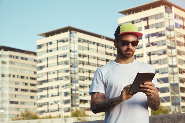 城镇一个严肃的年轻人穿着白色的普通t恤 戴着红黄黑相间的卡车司机帽 对着城市建筑和天空看着他的平板电脑电脑触摸屏电子
