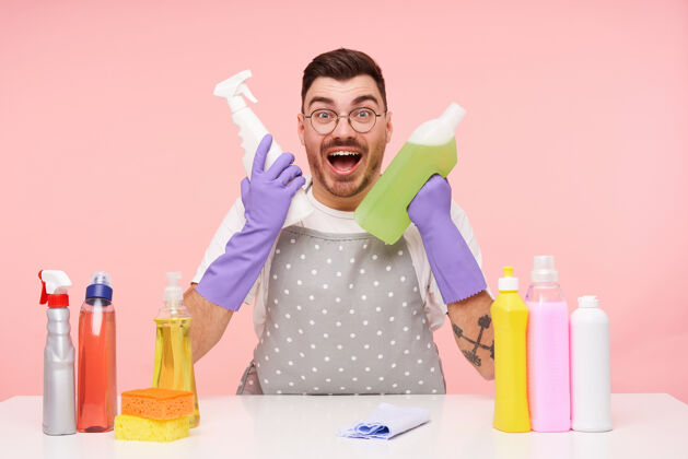 眼镜一个戴着眼镜的年轻的黑发男人兴高采烈地看着 举起手来拿着家用化学品 隔离在粉红色的皮肤上瓶子洗衣服表情