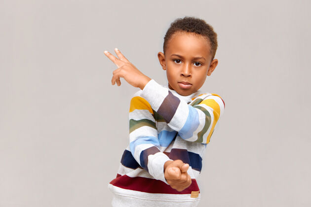 肖像一幅孤立的画面 自信英俊的非洲裔美国男孩穿着条纹套头衫 指着空白的墙壁 用手指画出两个手势 表情严肃地盯着右边非洲成功孩子