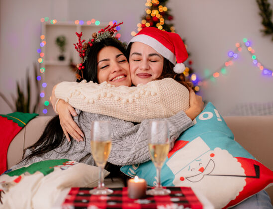 扶手椅戴着圣诞帽的漂亮姑娘们坐在扶手椅上互相拥抱 在家里享受圣诞时光年轻圣诞每个