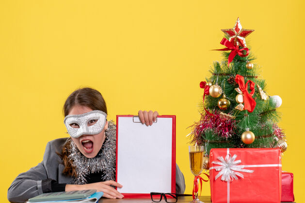 桌子正面图：戴着面具的年轻女孩坐在圣诞树和鸡尾酒礼物的桌边大喊大叫礼物树鸡尾酒