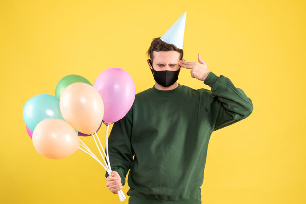 寺庙正面图：戴着派对帽和五颜六色气球的年轻人把手指枪放在他站在黄色的太阳穴上生日站着庆祝