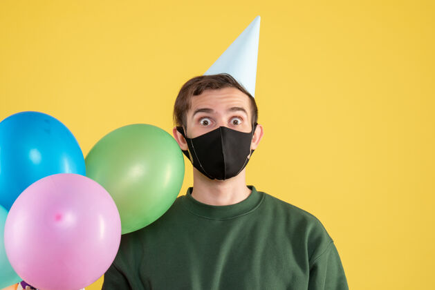 面具正面图：戴着派对帽和黑色面具的年轻人黄色的彩色气球肖像脸乐趣