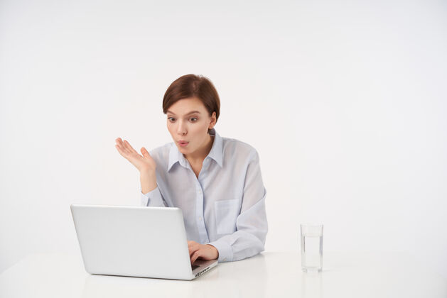 坐着惊讶的年轻可爱的短发黑发女子坐在白色的桌子上 用现代化的笔记本电脑 睁大眼睛看着屏幕 穿着正式的衣服情绪办公室员工