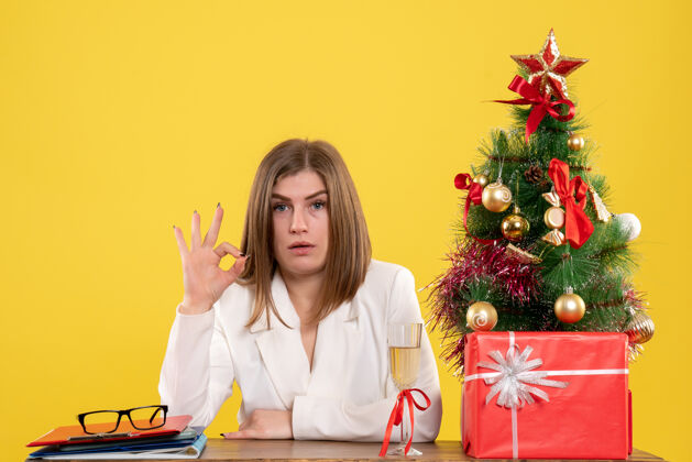 成人前视图：女医生坐在黄色地板上的桌子前 手里拿着圣诞树和礼品盒坐着女医生职业