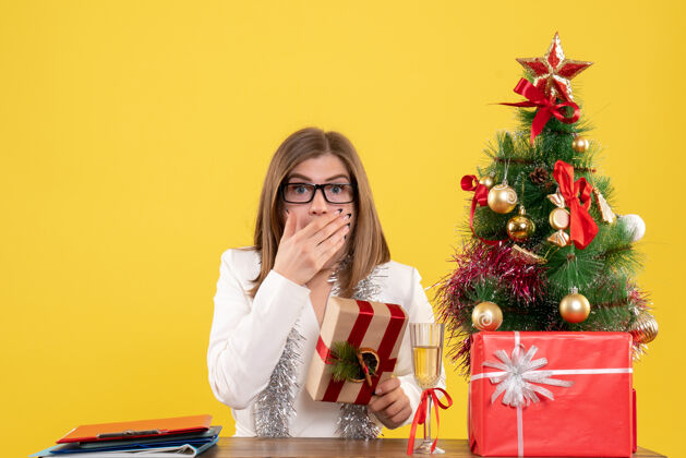 前面正面图：女医生坐在桌子前 手里拿着黄色的礼物 还有圣诞树和礼盒情感成人微笑
