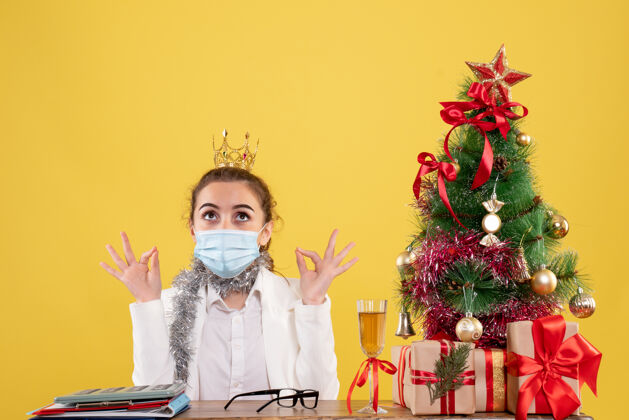 肖像正面图女医生戴着黄色背景的无菌口罩坐在圣诞树和礼盒里快乐人女医生