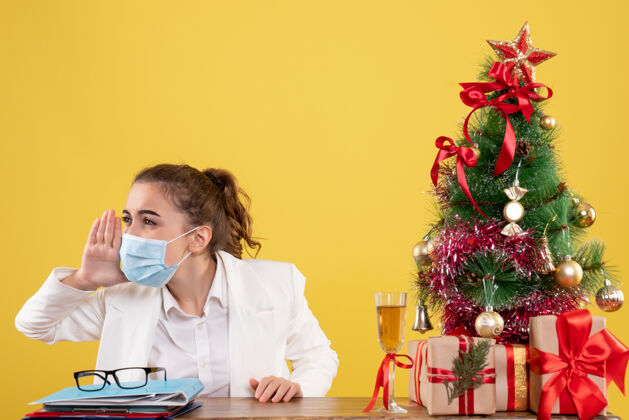 面具正面图女医生戴着无菌口罩坐在黄色背景上呼唤圣诞树和礼品盒人女性电话