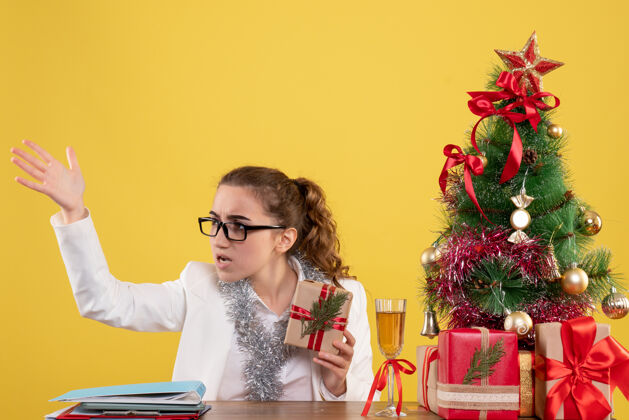 漂亮前视图：女医生拿着圣诞礼物和黄色背景上的圣诞树坐着女性办公室快乐
