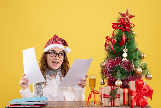抱着前视图女医生坐在圣诞树上 拿着黄色背景上的文件坐着情感礼物