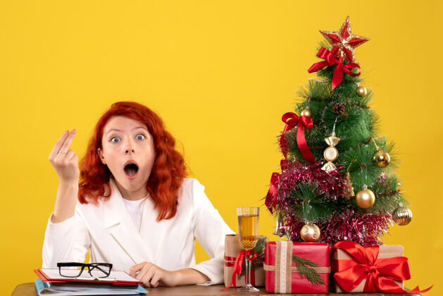 女医生正面图：女医生坐在桌子后面 拿着圣诞礼物 黄色背景上的树健康坐着前面