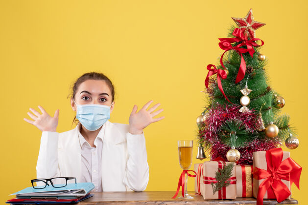专业正面图女医生戴着黄色背景的无菌口罩坐在圣诞树和礼盒里医学前面面具