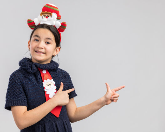 编织小女孩穿着针织连衣裙 头上系着一条红色领带 戴着一个有趣的圣诞戒指 看起来很开心 积极地用食指指着旁边快乐积极手指