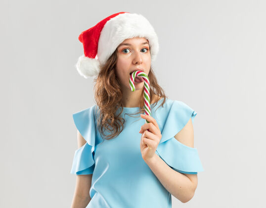 快乐穿着蓝色上衣 戴着圣诞帽 手持糖果手杖的年轻女子快乐地去品尝它新味道年轻