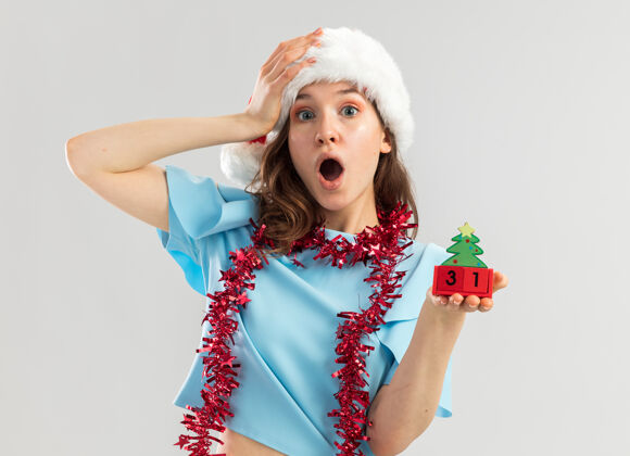 惊奇身穿蓝色上衣 脖子上戴着镀金圣诞帽的年轻女子手里拿着玩具方块 手放在头上 看起来很惊奇看帽子顶部