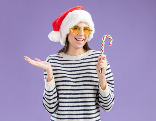 快乐戴着太阳眼镜 戴着圣诞帽 面带微笑的白人女孩手持糖果手杖 手一直张开糖果复制圣诞