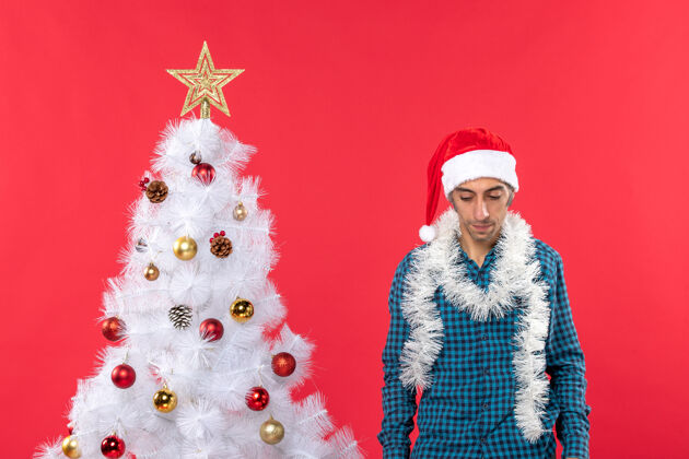 站着一个戴着圣诞老人帽子 穿着蓝色条纹衬衫 站在红色圣诞树旁低头的悲伤的年轻人礼物衬衫年轻
