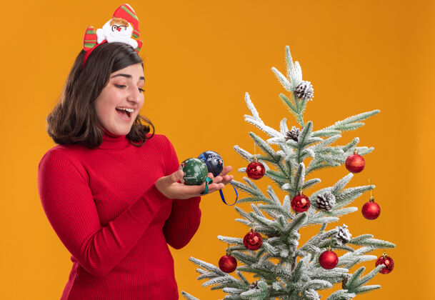 圣诞节穿着圣诞毛衣的快乐小女孩戴着有趣的头带拿着圣诞球在橙色背景下的圣诞树旁圣诞树女孩头带
