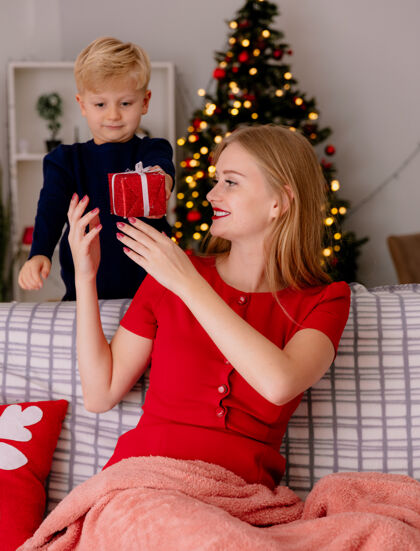 站着在一间背景是圣诞树的装饰房间里 穿着红色连衣裙的快乐妈妈坐在沙发上微笑着 她的小孩站在后面给妈妈送礼物装饰衣服微笑