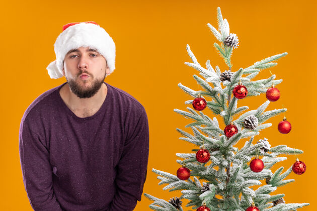 无聊穿着紫色毛衣 戴着圣诞帽的年轻人站在橙色背景下的圣诞树旁 看上去又累又无聊站着圣诞树圣诞老人
