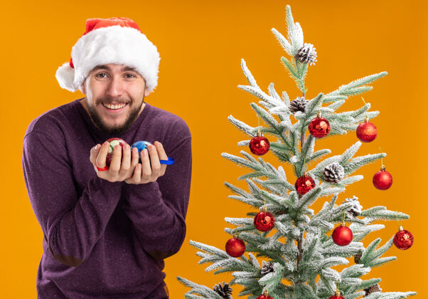 帽子身穿紫色毛衣 戴着圣诞帽 手持圣诞球的年轻人站在橙色背景下的圣诞树旁 开心而兴奋地微笑着站着兴奋微笑
