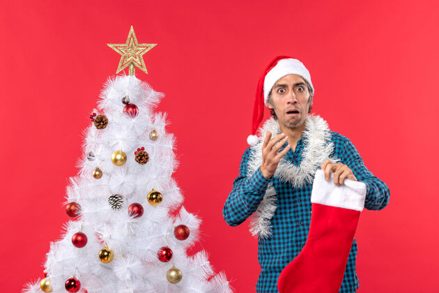 衬衫困惑的年轻人戴着圣诞老人的帽子 穿着蓝色的衬衫 手里拿着圣诞礼物男人庆祝圣诞老人
