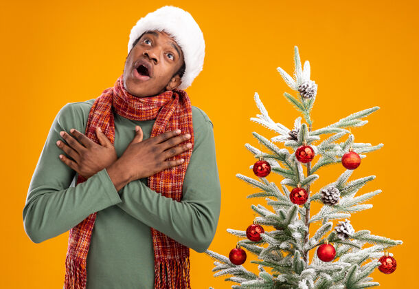 惊讶戴着圣诞帽 围着围巾的非洲裔美国人抬头望着 手放在胸前 在橙色背景下的圣诞树旁惊喜不已美国人男人围着