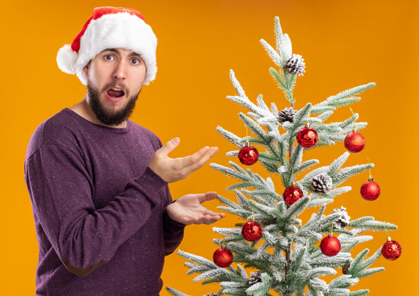 介绍穿着紫色毛衣 戴着圣诞帽的年轻人站在橙色背景下的圣诞树旁 看上去既惊讶又惊讶毛衣男人圣诞树