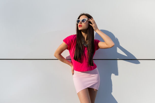 时尚穿着夏装戴着太阳镜的时髦女人在白色的城市墙上摆姿势可爱女孩成人