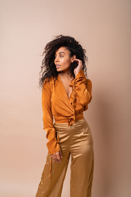 女主角自信的非洲女人 一头完美的卷发 穿着休闲的橙色上衣和金色裤子 在米色的墙上摆着姿势性感化妆魅力
