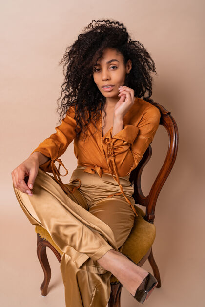 奢华时尚年轻诱人的非洲模特 完美的卷发 优雅的橙色衬衫和丝绸裤子 坐在老式椅子米色墙上模特诱惑晒黑
