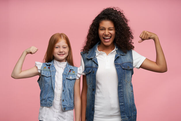 深色两位快乐的年轻女士站在粉红色的背景下 举起双手 高兴地展示她们强壮的二头肌 穿着牛仔裤背心和白衬衫情绪狐狸心情