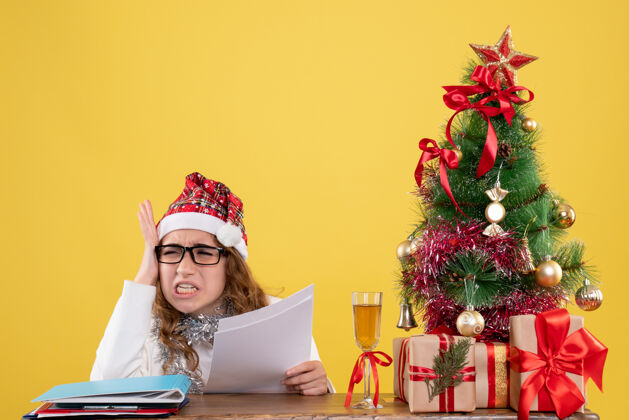 礼物前视图女医生坐在圣诞树上 拿着黄色背景上的文件抱着礼物文件