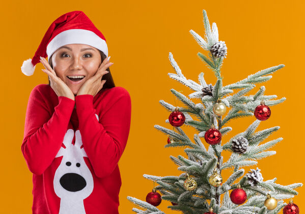 女孩惊讶的年轻亚洲女孩戴着圣诞帽 穿着毛衣站在圣诞树旁 嘴巴被遮住 手被隔离在橙色的背景上树亚洲人年轻
